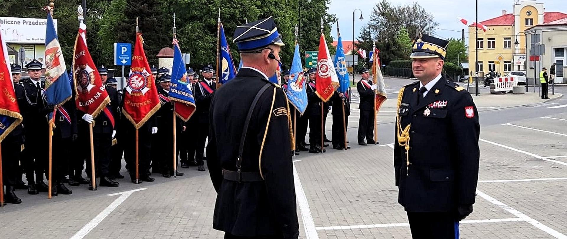 Dwóch oficerów PSP stojących naprzeciw siebie. Jeden z nich jest w stopniu generała. W tle, ustawieni w szeregu strażacy. Część z nich trzyma sztandary jednostek.