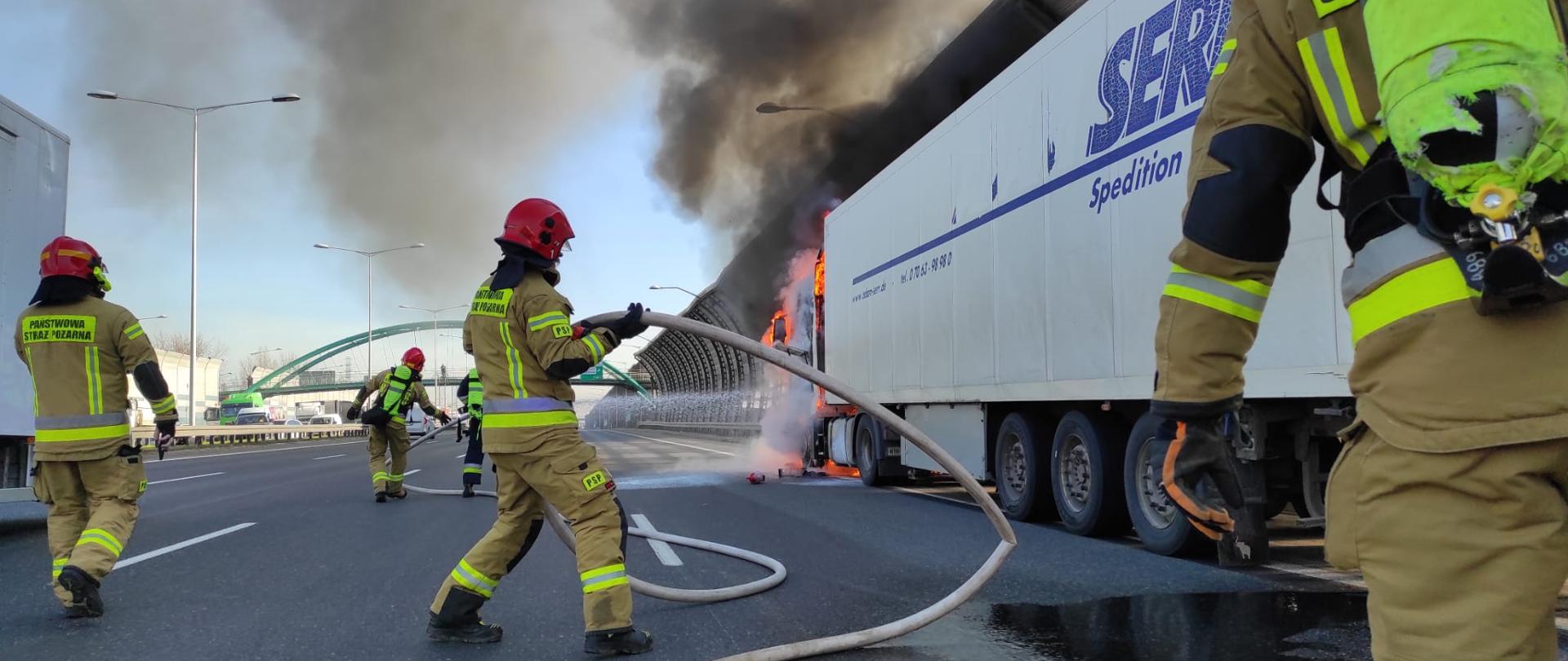 Droga szybkiego ruchu na której poboczu stoi samochód ciężarowy. Z samochodu wydobywają się płomienie i czarny gęsty dym. Strażacy rozwijający węże potrzebne do ugaszenia pożaru.
