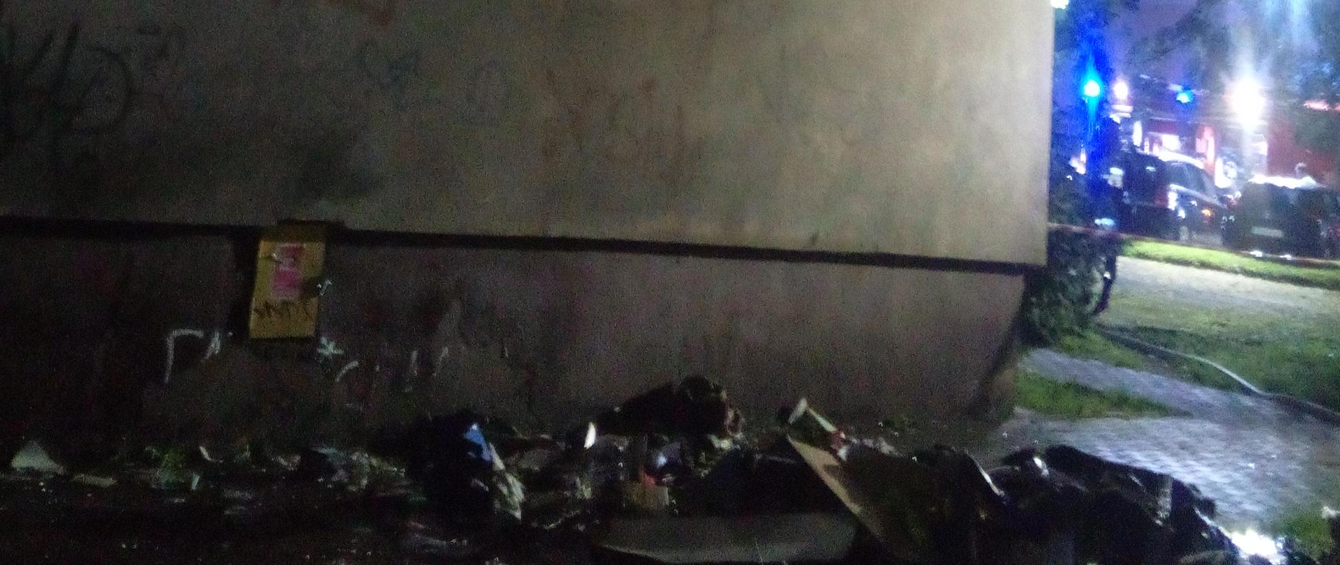 Zdjęcie przedstawia elewację bloku 4 piętrowego. Na dole widać pozostałości po pożarze wyrzucone przez okno. W tle znajduje się samochód straży pożarnej
