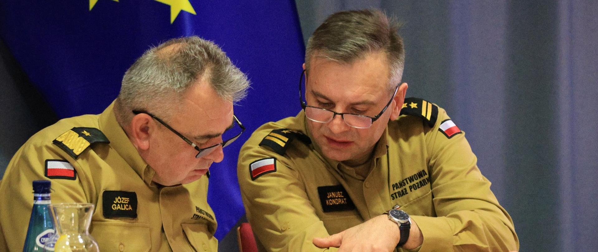 Na zdjęciu dwóch wysokich rangą funkcjonariuszy Państwowej Straży Pożarnej w beżowych mundurach analizuje dokumenty podczas oficjalnego spotkania. Nadbrygadier Józef Galica oraz Janusz Kondrat pochylają się nad otwartą teczką, wskazując na konkretne fragmenty tekstu. W tle widoczna jest flaga Unii Europejskiej. Na stole stoją szklana karafka z wodą i cytryną oraz butelka wody mineralnej.