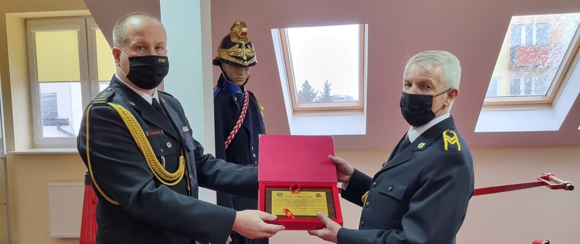 Zdjęcie przedstawia dwóch strażaków w mundurach galowych. Jeden z nich- starszy brygadier Paweł Jakubowski (Komendant Powiatowy PSP w Jędrzejowie) wręcza strażakowi odchodzącemu na zaopatrzenie emerytalne grawerton gratulacyjny. Strażacy znajdują się na sali tradycji. W tle widnieje manekin strażaka ubranego w zabytkowy mundur galowy. 