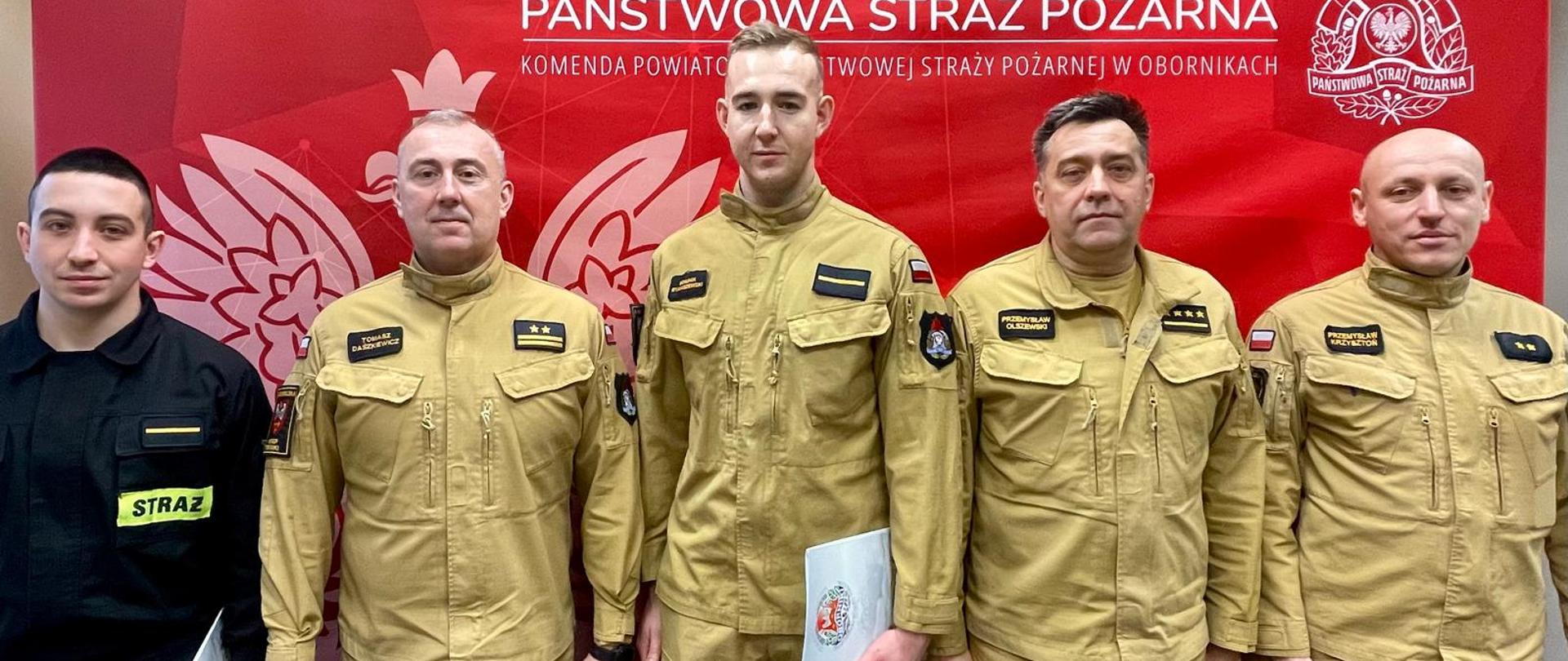 Grupowe zdjęcie sześciu strażaków stojących obok siebie w umundurowaniu służbowym. Pięciu mężczyzn ma jasnobeżowe mundury Państwowej Straży Pożarnej, jeden po lewej stronie ma czarny strój z napisem „STRAŻ”. W tle czerwona ścianka z białym godłem i napisem „Państwowa Straż Pożarna”. Kilku strażaków trzyma dokumenty; wszyscy stoją frontalnie, pozując do zdjęcia.