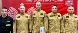 Grupowe zdjęcie sześciu strażaków stojących obok siebie w umundurowaniu służbowym. Pięciu mężczyzn ma jasnobeżowe mundury Państwowej Straży Pożarnej, jeden po lewej stronie ma czarny strój z napisem „STRAŻ”. W tle czerwona ścianka z białym godłem i napisem „Państwowa Straż Pożarna”. Kilku strażaków trzyma dokumenty; wszyscy stoją frontalnie, pozując do zdjęcia.