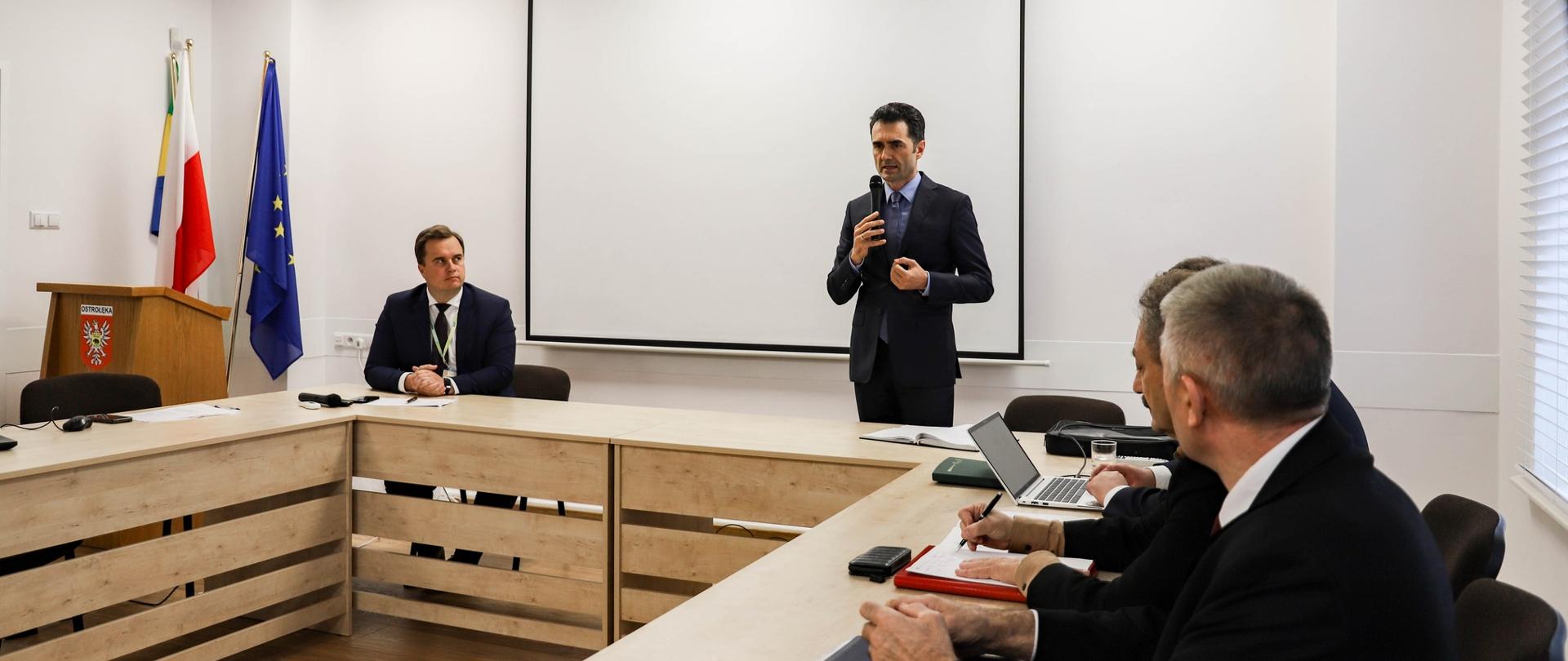 Na zdjęciu widać stojącego mężczyznę z mikrofonem oraz trzech siedzących przy biurkach