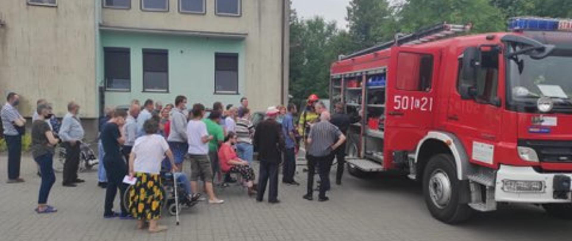Wóz strażacki zaparkowany przed szarym budynkiem. Strażacy prezentują wyposażenie zgromadzonym osobą 