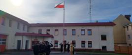 Uroczystość z okazji Dnia Flagi Rzeczypospolitej Polskiej. Strażacy ze zmian służbowych stoją naprzeciwko siebie. Poczet flagowy wciąga flagę państwową na maszt. Komendant, Zastępca Komendanta, poczet flagowy i dowódcy zmian salutują.