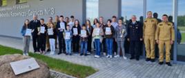 Zdjęcie przedstawia uczestników konkursu trzymających dyplomy, medale oraz puchary, opiekunów oraz organizatorów konkursu podczas pamiątkowej fotografii na zakończenie ogólnopolskiego konkursu wiedzy pożarniczej. Zdjęcie zrobione jest przed budynkiem Jednostki Ratowniczo-Gaśniczej nr 3 w Rzeszowie.
