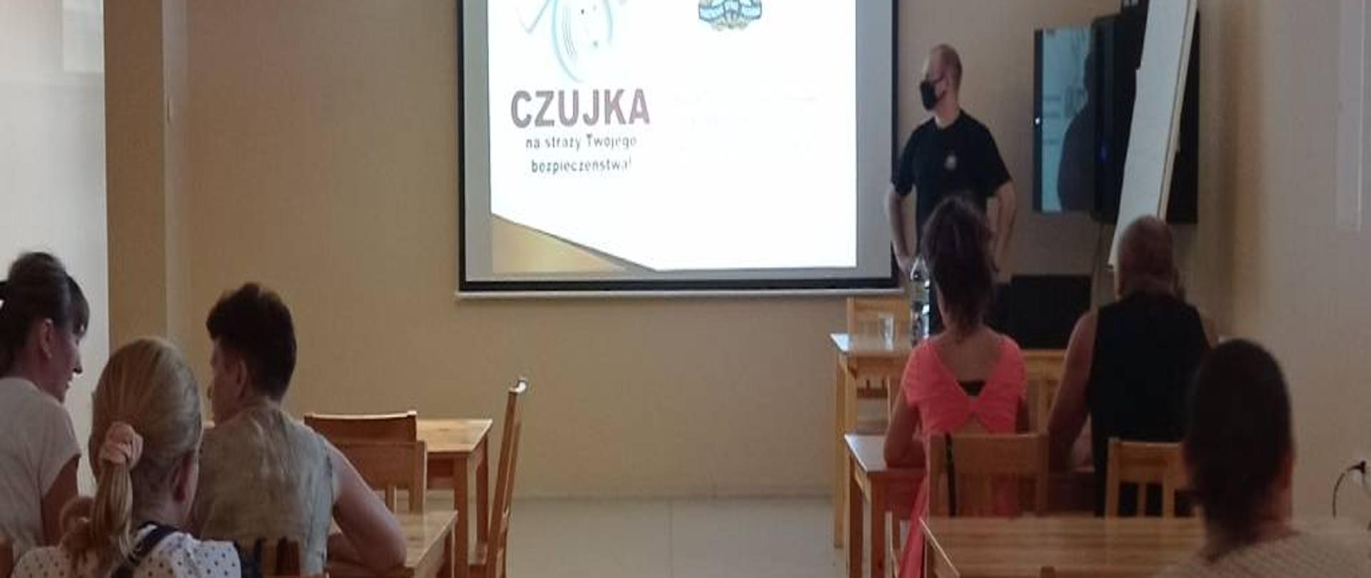 Zdjęcie przedstawia uczestników programu organizowanego przez Centrum Integracji Społecznej w Zambrowie siedzących w ławkach podczas spotkania profilaktycznego z funkcjonariuszem Komendy Powiatowej Państwowej Straży Pożarnej w Zambrowie. Omawiający prezentację funkcjonariusz ubrany w krótką czarną koszulkę z logiem PSP oraz spodnie z ubrania dowódczo-sztabowego z pasem. Zdjęcie wykonano w jednym z pomieszczeń wykładowych znajdujących się w budynku Bursy Szkolnej nr 1.