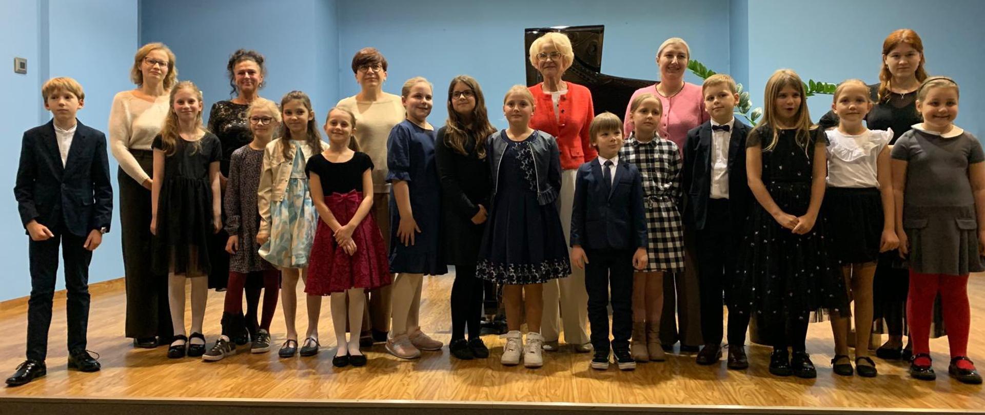 Na zdjęciu widać grupę osób stojących na scenie w sali o niebieskich ścianach i jasnym suficie z oświetleniem punktowym. W pierwszym rzędzie stoi kilkanaścioro dzieci, ubranych elegancko — dziewczynki w sukienkach, chłopcy w garniturach. Za nimi stoi nauczycielki, również ubrane odświętnie. W tle widać fortepian. Cała grupa pozuje do wspólnego zdjęcia, uśmiechając się w stronę aparatu.