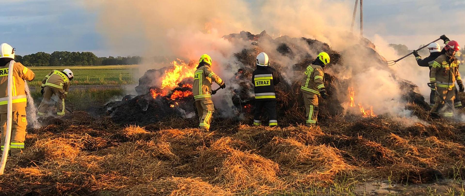 Widoczni strażacy gaszący płonący stóg słomy.