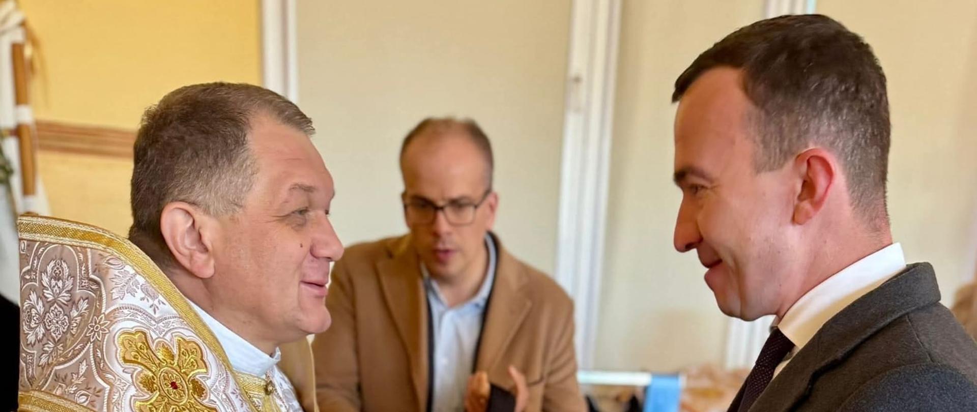 Zdjęcie przedstawia trzech mężczyzn, jeden wręcza drugiemu podarunek