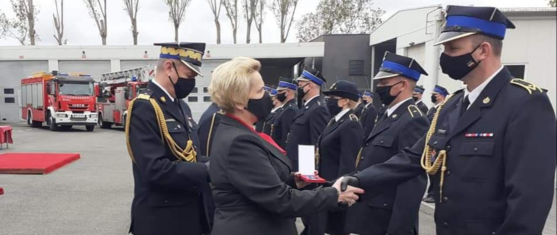 Na zdjęciu na pierwszym planie 3 osoby. Po lewej stronie stojąca ubrana na biało-czarno poseł Porowska. Ustawiona bokiem do kadru, ma ciemne długie włosy, sznur pereł na szyi. Po prawej stronie i zarazem centralnie w kadrze wicewojewoda ubrana w czerwoną sukienkę i czarny żakiet. Ściska dłoń jednemu ze strażaków, który stoi na przeciw niej. W tle pozostali strażacy. 