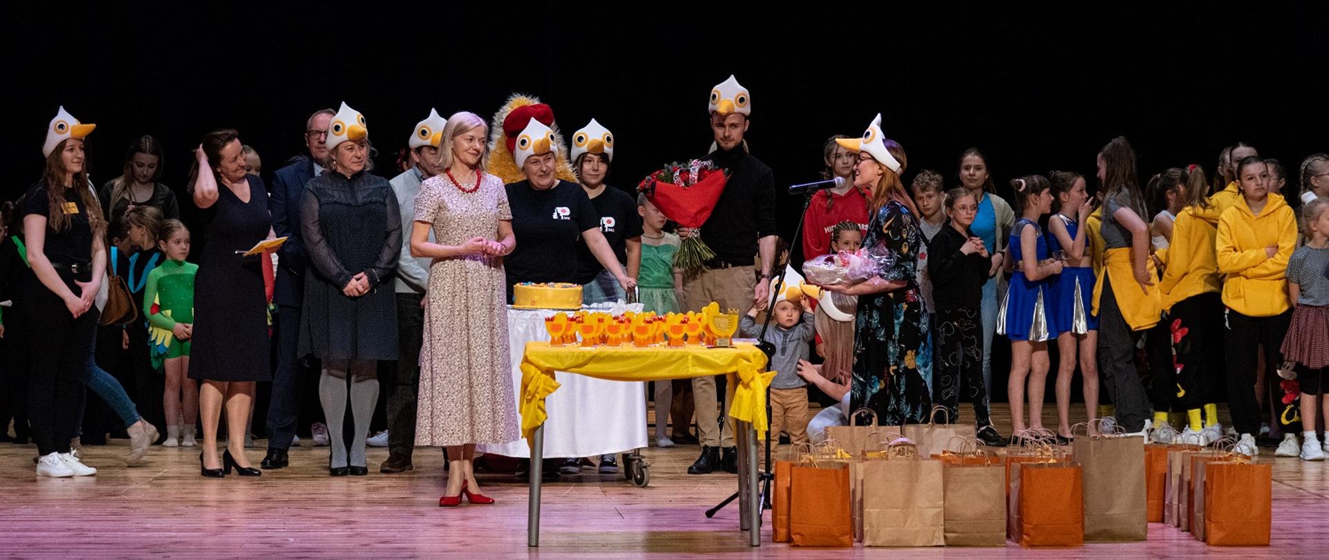 Na scenie stoją organizatorzy oraz uczestnicy Przeglądu "Kaczucha". Na pierwszym planie stolik a na nim szklane żółte statuetki i nagrody dla uczestników. 