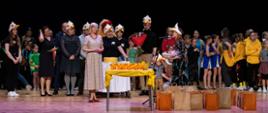 Na scenie stoją organizatorzy oraz uczestnicy Przeglądu "Kaczucha". Na pierwszym planie stolik a na nim szklane żółte statuetki i nagrody dla uczestników. 