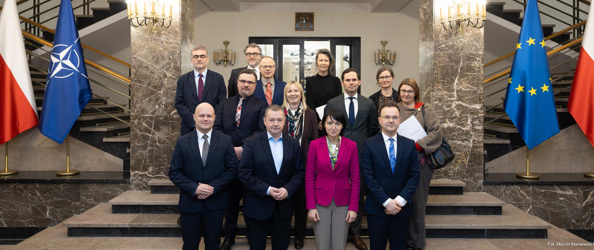 Na marmurowych schodach stoi grupa osób ustawiona w trzech rzędach. Po bokach stoją po 2 flagi.