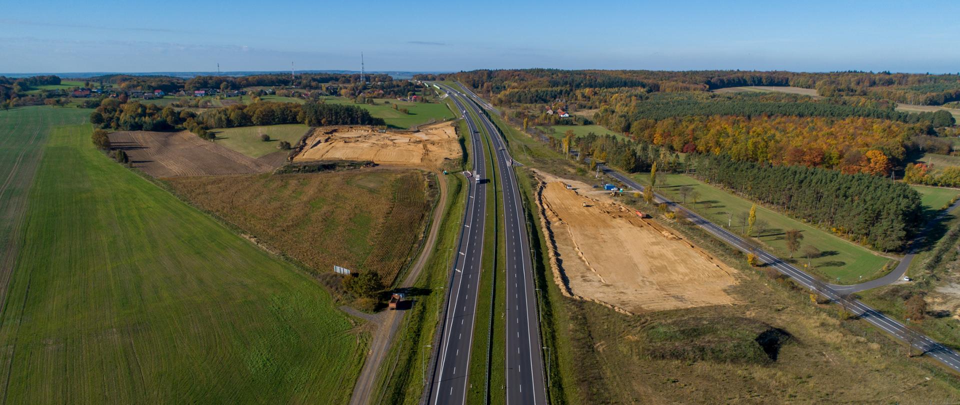 Droga dwujezdniowa otoczona polami i lasami. Po obu stronach drogi teren przygotowany pod budowę Miejsc Obsługi Podróżnych.