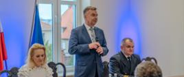 Minister Wieczorek stoi nad stołem i mówi, obok niego siedzą mężczyzna w szarym mundurze i kobieta w białej bluzce, naprzeciwko na ustawionych w rzędy krzesłach siedzą ludzie.