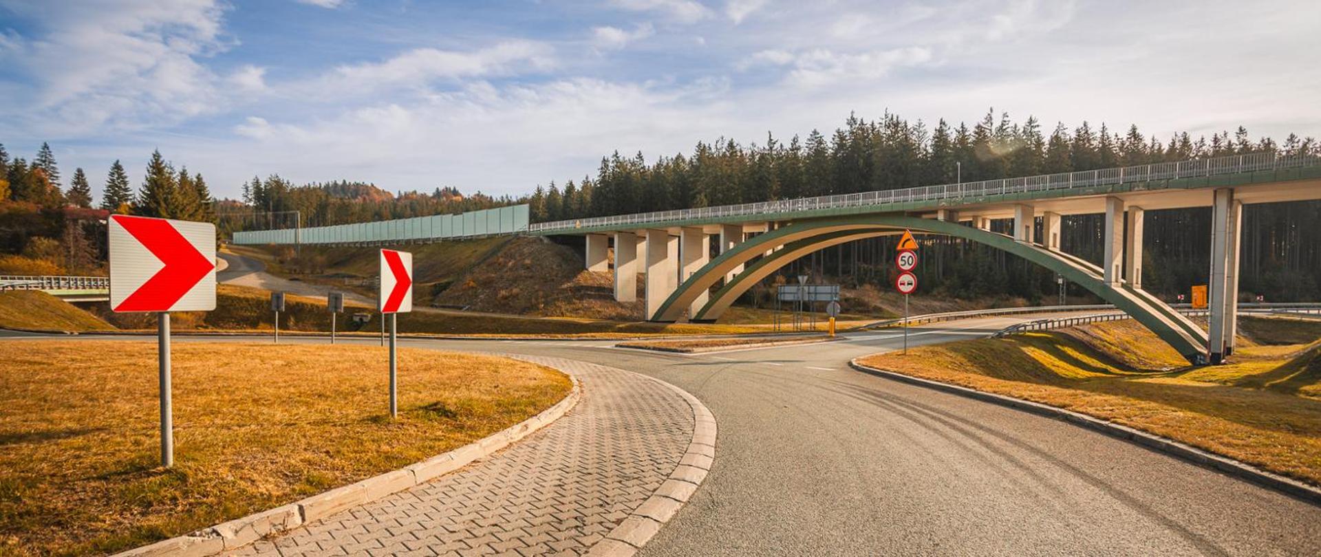 Most drogowy nad jezdnią, obok skrzyżowania. Na pierwszym planie znaki drogowe, a w tle las i ekrany akustyczne.