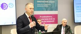 Minister Jacek Żalek przemawia na konferencji, na monitorze na pierwszym planie nazwa konferencji „Jak skutecznie tworzyć strategie B+R w polskim przemyśle?”, w tle ekran o banner z hasłami wydarzenia oraz inni prelegenci na fotelach
