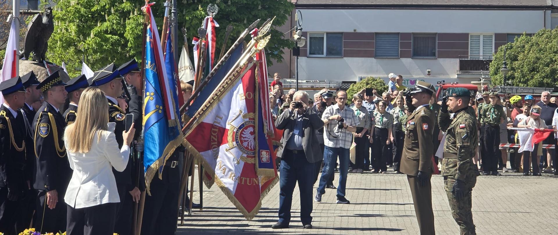 Uczestnicy i mundury
• Wojsko Polskie: Po prawej stronie widać dwóch żołnierzy oddających honor przez salutowanie. Jeden z nich ubrany jest w oliwkowy mundur galowy i rogatywkę, drugi w mundur polowy i zielony beret.
• Służby mundurowe: Po lewej stronie stoją funkcjonariusze w ciemnogranatowych mundurach galowych ze złotymi akselbantami (sznurami), prawdopodobnie przedstawiciele Straży Pożarnej.
• Poczty sztandarowe: Centralną część zdjęcia zajmują liczne poczty sztandarowe. Wyraźnie widać sztandar z napisem "BÓG HONOR OJCZYZNA" oraz emblematem Związku Ochotniczych Straży Pożarnych RP.
• Harcerze i cywile: W tle, za barierkami, zgromadzony jest tłum mieszkańców, wśród których widać harcerzy w szarych i zielonych mundurach.
Szczegóły i kompozycja
• Dokumentacja wydarzenia: Na zdjęciu widać osoby fotografujące uroczystość – kobietę w białej marynarce na pierwszym planie po lewej oraz mężczyznę z profesjonalnym aparatem w centrum placu.
• Otoczenie: Akcja dzieje się na wybrukowanym placu miejskim. W tle widoczne są bloki mieszkalne oraz zieleń miejska (kasztanowce). Po lewej stronie fragment pomnika z orłem, który był lepiej widoczny na poprzednich zdjęciach (zdjecie 1.jpg i zdjecie 2.jpg).
Zdjęcie dobrze oddaje podniosłą atmosferę oficjalnych obchodów, łączącą przedstawicieli wojska, służb ratowniczych, młodzieży oraz lokalnej społeczności.
