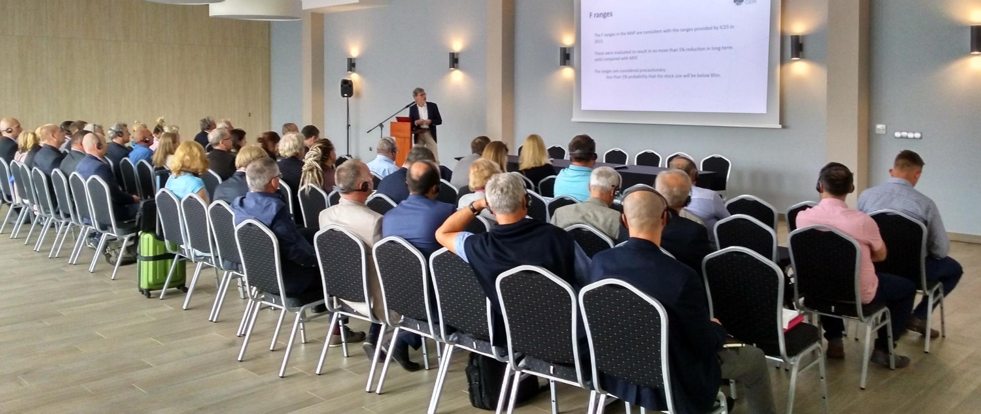 Uczestnicy konferencji słuchają prelegenta. Na rzutniku widać prezentację multimedialną.