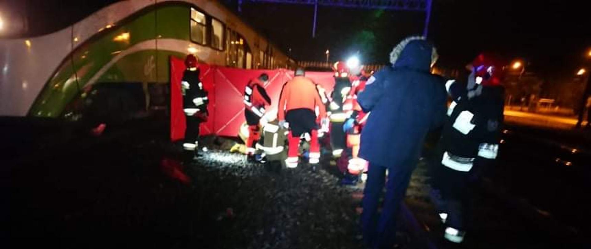 Na zdjęciu widać strażaków i ratowników medycznych udzielających pierwszej pomocy. Teren akcji jest oświetlony. Poszkodowany osłonięty parawanem, na drugim planie widać pociąg osobowy