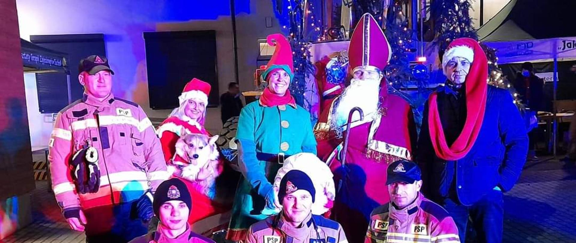 Zdjęcie przedstawia grupę ośmiu osób. Wśród nich jest 4 strażaków w ubraniach specjalnych. Obok strażaków stoją osoby w świątecznych przebraniach: Święty Mikołaj, elf. Pani Mikołajowa trzyma na rękach psa. W tle widać budynek i gałązki choinki przyozdobione lampkami. 