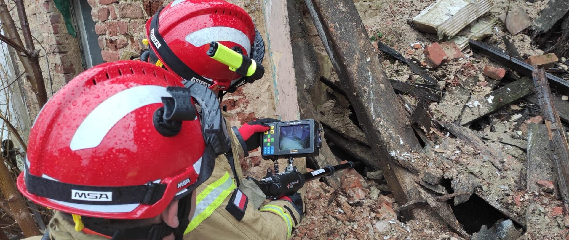 Strażacy ze Specjalistycznej Grupy Poszukiwawczo-Ratowniczej "Brzeg" przy pomocy kamery wziernikowej wykluczają obecność osób poszkodowanych pod gruzami. W tle widoczna część zawalonego obiektu. Strażacy w ubraniach specjalnych i kaskach koloru czerwonego.
