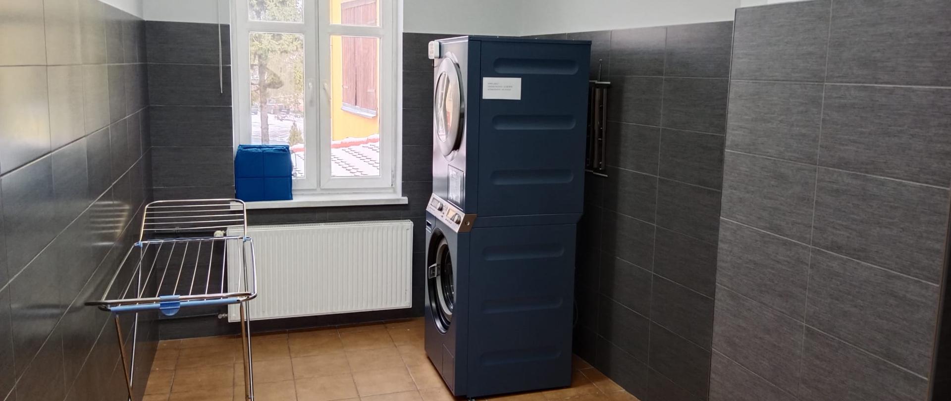 Widok pna pomieszczenie gospodarcze. Na wprost okno. Po prawej stronie pomieszczenia pralka a na niej suszarka. Po lewej suszarka na ubrania.