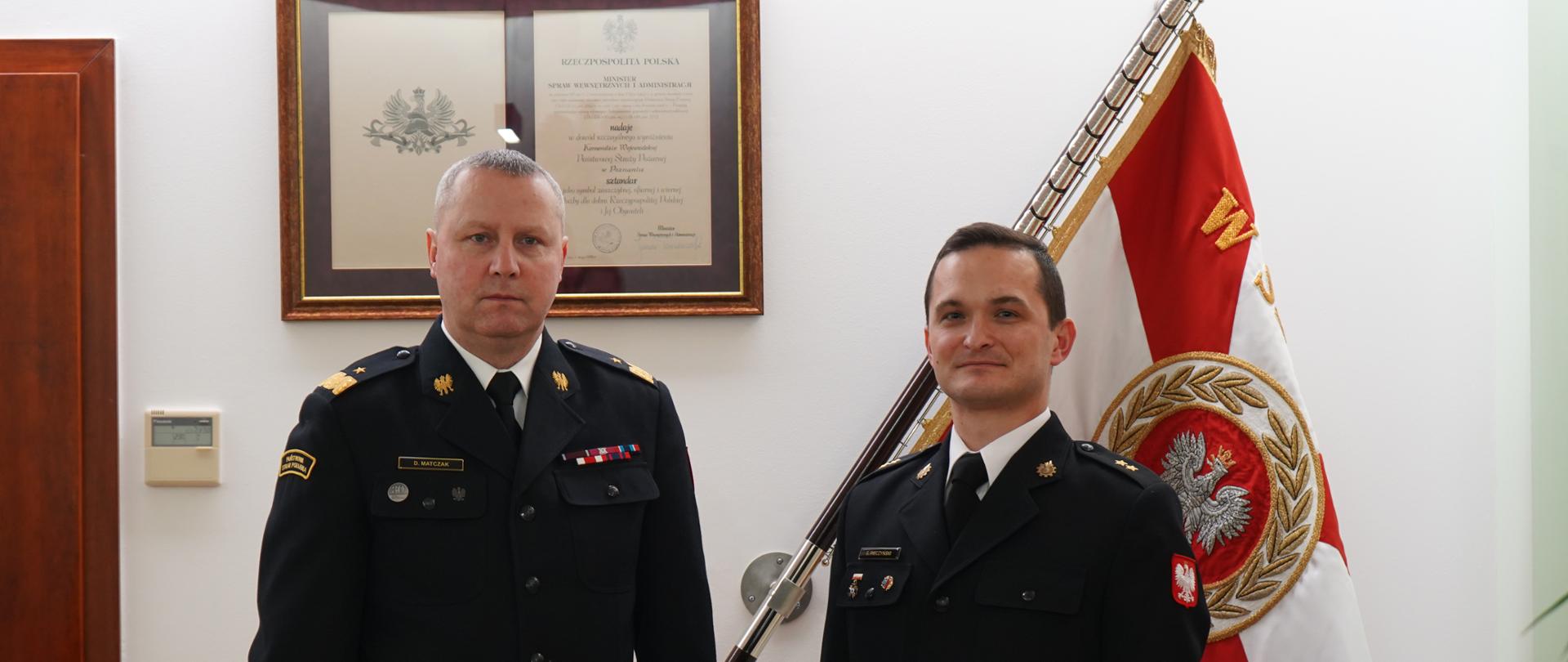 dwóch strażaków w ciemnych mundurach białych koszulach z krawatem stoi obok siebie, jeden z nich trzyma czerwoną teczkę w ręku, w tle sztandar
