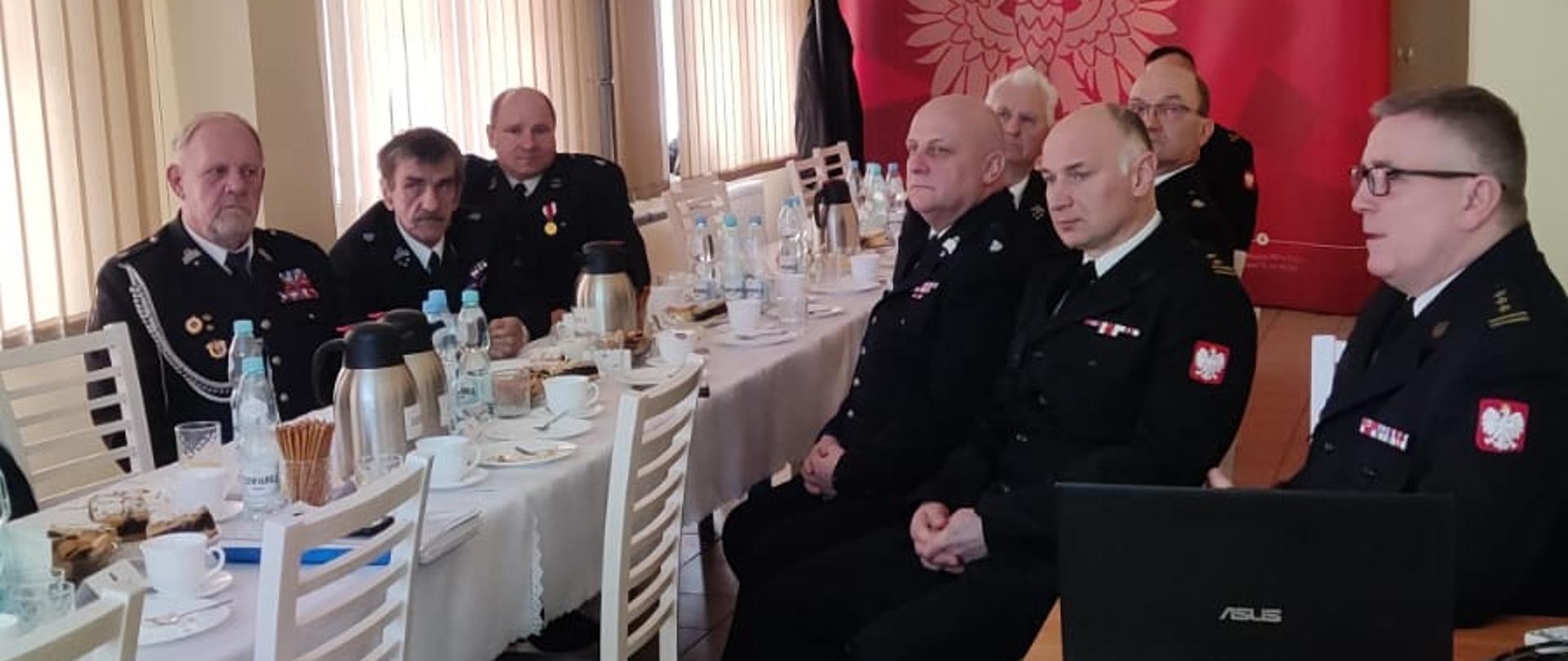 Strażacy w mundurach wyjściowych siedzą przy stole nakrytym białym obrusem. Na stole dzbanki z kawą i herbatą. W tle ścianka firmowa Państwowej Straży Pożarnej.