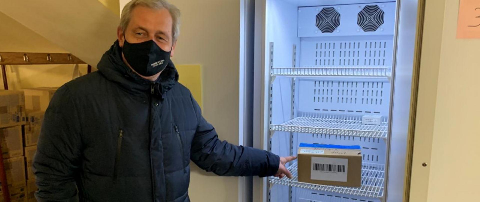 Wojewoda stoi obok lodówki z otwartymi drzwiczkami. w lodówce znajduje się mały karton - a w nim fiolki ze szczepionkami przeciwko koronawirusowi. Wojewoda wskazuje palcem na pudełko. Wojewoda ubrany w ciemną, zimową kurtkę.