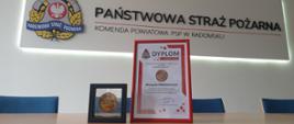 Na pierwszym planie ustawione na stole odznaka oraz dyplom w ramkach. W tle logo psp oraz napis Państwowa Straż Pożarna Komenda Powiatowa PSP w Radomsku