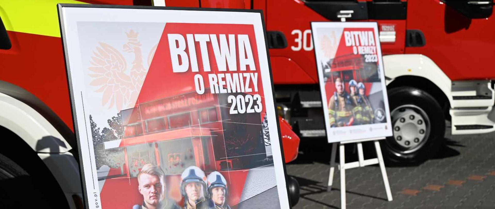Zdjęcie przedstawia plakat akcji "Bitwa o remizy 2023" w antyramie na sztaludze. W tle czerwone strażackie samochody oraz drugi plakat na sztaludze.