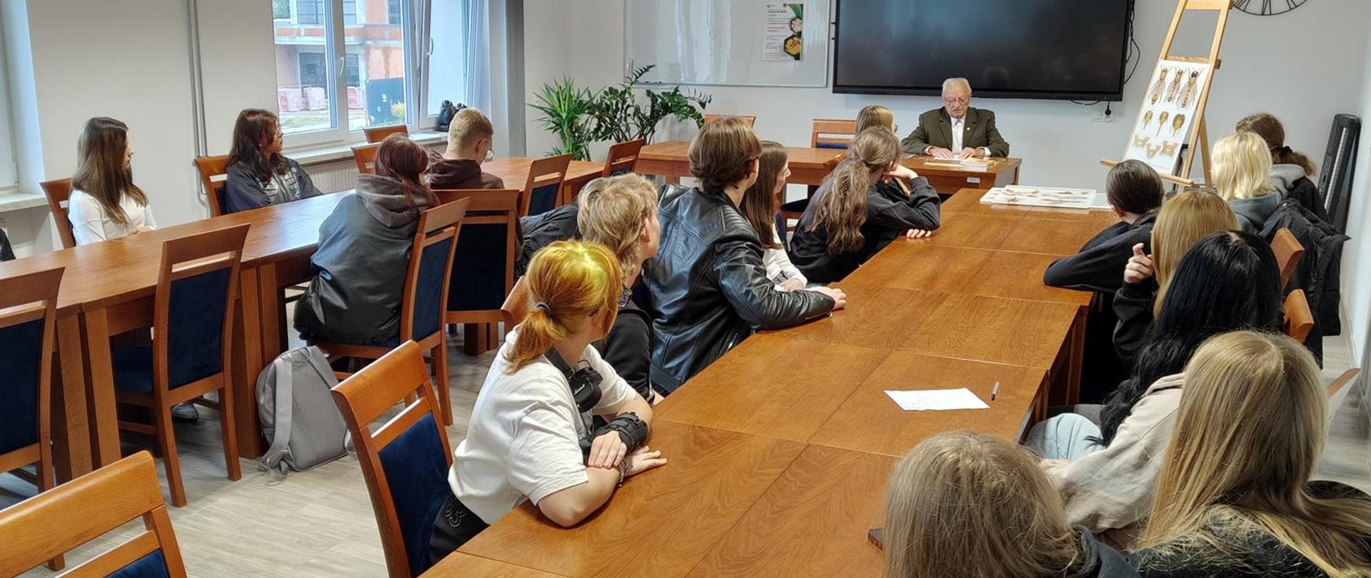 Starszy mężczyzna czyta, siedząc przy stole. Słuchają go uczniowie.