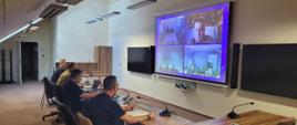 Uczestnicy wideokonferencji przy stole w KG PSP i pozostali widoczni na monitorze