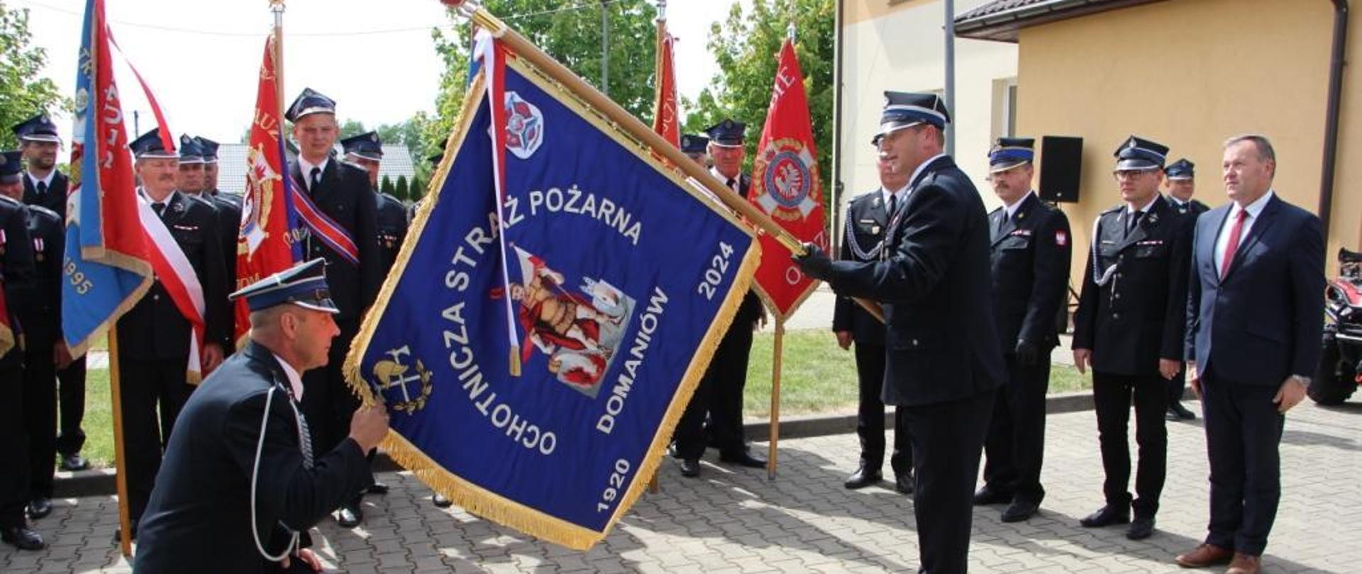 Zdjęcie wykonane podczas uroczystości z okazji nadania sztandaru oraz przekazania pojazdu typu quad dla jednostki Ochotniczej Straży Pożarnej w Domaniowie. Na zdjęciu przed jednostką widać ustawionych od lewej w dwóch szeregach jeden za drugim członów jednostki w Domaniowie. Dalej za nimi ustawione są poczty sztandarowe z pozostałych jednostek z terenu gminy. Strażacy ubrani są w mundury galowe w kolorze ciemnym granatowym. Przed nimi następuje uroczysty moment przekazania sztandaru dla jednostki. Prezes jednostki klęczy na jednym kolanie przed pochylonym sztandarem. Dodatkowo jedną ręką trzyma za róg sztandaru. Na przeciwko niego stoi prezes zarządu oddziału powiatowego który pochyla sztandar przed klęczącym strażakiem. Za nimi w tle widać przedstawiciela Państwowej Straży Pożarnej oraz przedstawiciel władz samorządowych z terenu gminy. 