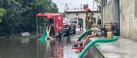 Strażacy w akcji na zalanym terenie, używający pomp do wypompowywania wody. W tle widać czerwony pojazd strażacki z otwartą przyczepą oraz innych strażaków.