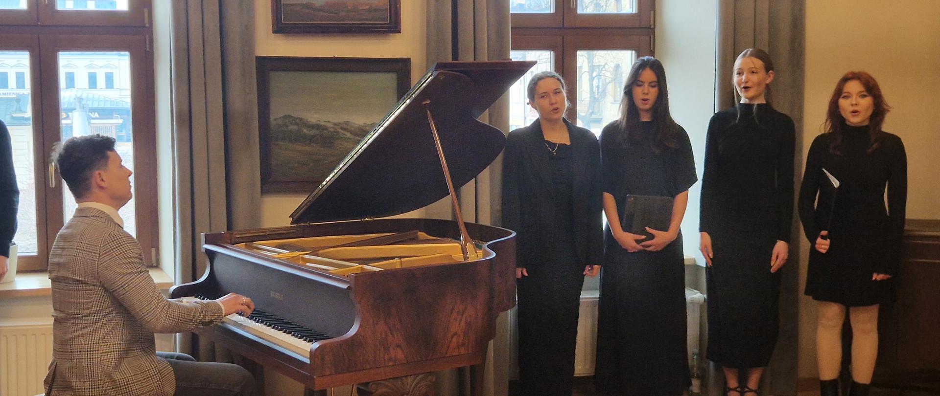 Zdjęcie przedstawia kameralny występ muzyczny w eleganckim, klasycznym wnętrzu. Po lewej stronie widać mężczyznę w jasnej marynarce w kratę, który siedzi przy otwartym fortepianie i gra na nim. Po prawej stronie stoi grupa czterech uczennic ubranych w eleganckie, czarne stroje, które wspólnie śpiewają. Centralnym punktem jest duży, brązowy fortepian o wysokim połysku z podniesioną klapą. Pomieszczenie ma drewnianą podłogę ułożoną w jodełkę. Na ścianie wiszą dwa obrazy w ramach, a duże okna z ciemnymi zasłonami wpuszczają do środka naturalne światło. Całość emanuje spokojną, uroczystą atmosferą, sugerującą koncert lub próbę w szkole muzycznej bądź instytucji kultury.