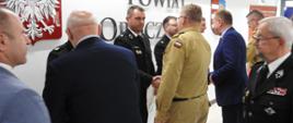 Na tle jasnej ściany stoją strażacy w ciemnych mundurach. Przed nimi mężczyźni i strażacy w jasnych mundurach. wszyscy podają sobie ręce. Za strażakami na ścianie widać napis Powiat oraz Godło Polski i herb powiatu tomaszowskiego. 