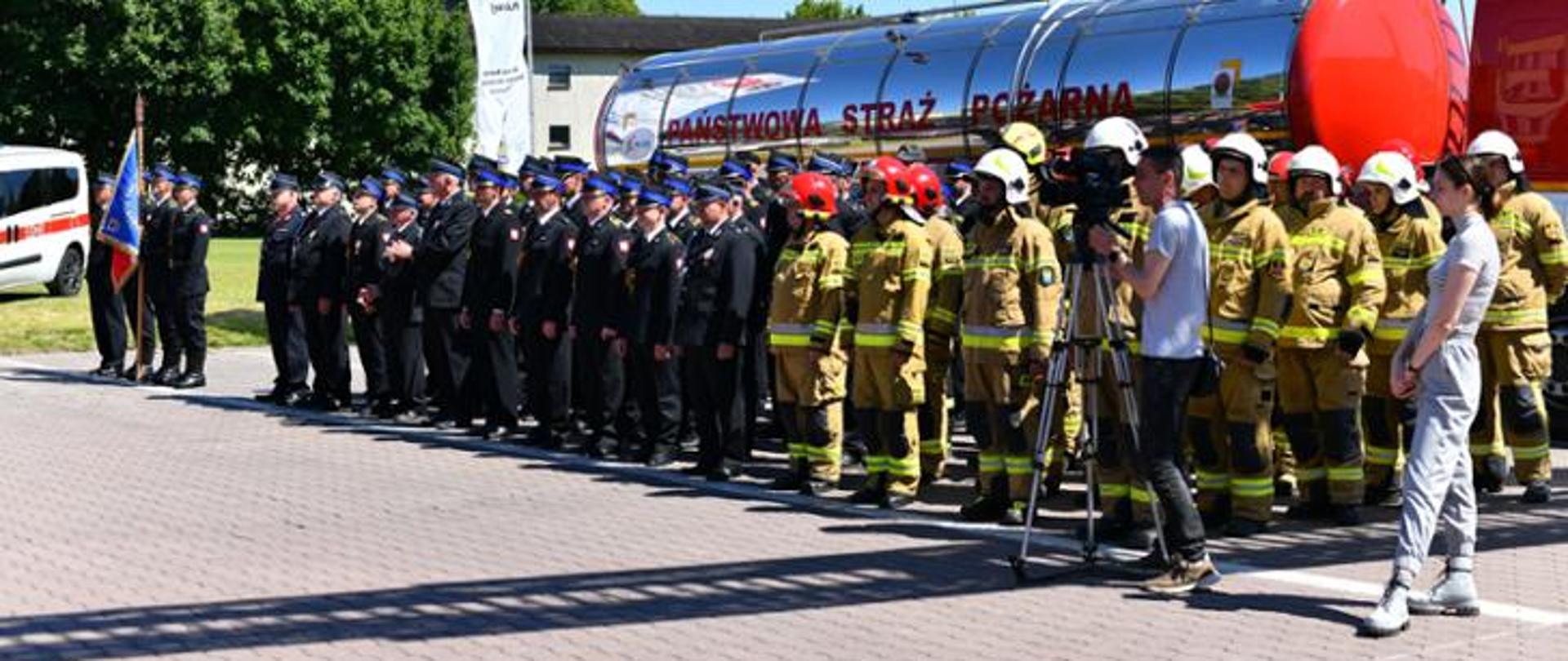Pododdziały strażaków w szyku, w tle cysterna z napisem PSP.
Widoczny poczet sztandarowy