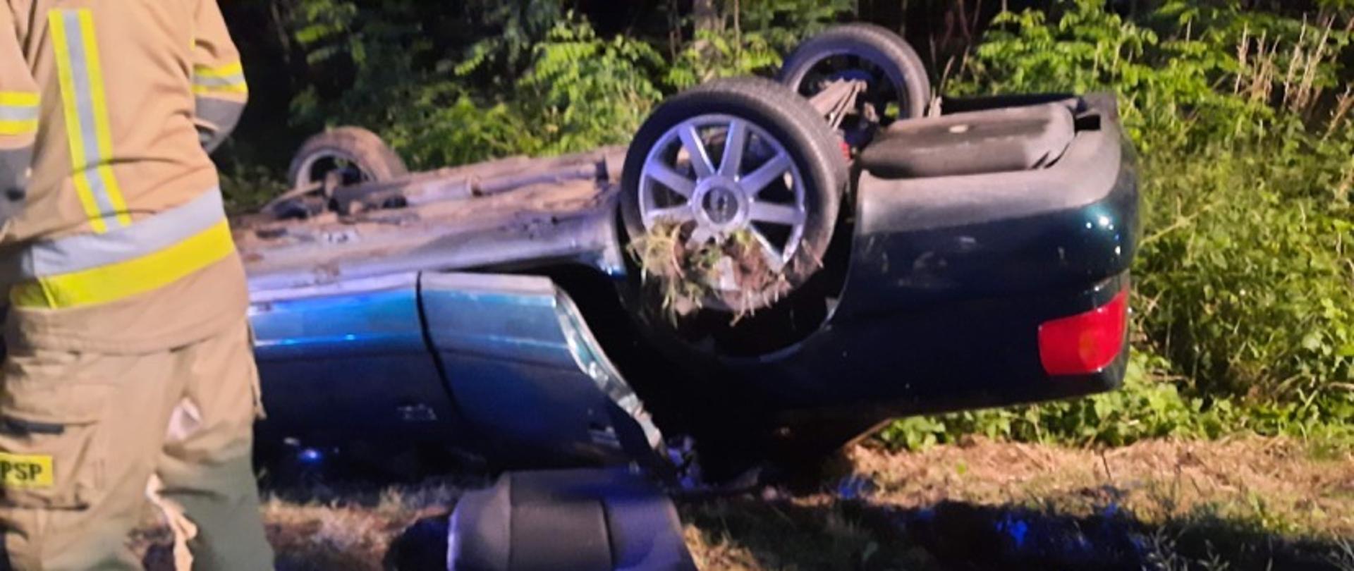 Samochód osobowy ciemnego koloru leży na dachu w przydrożnym rowie. Po lewej stronie stoi strażak