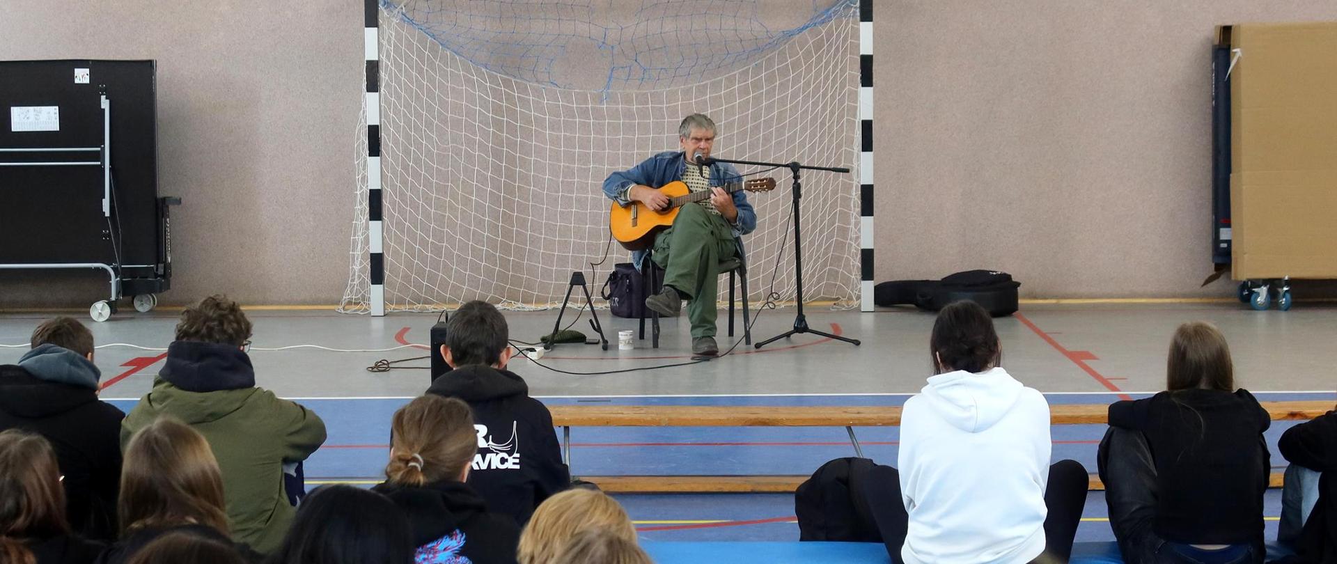 Sala gimnastyczna. Starszy mężczyzna gra na gitarze i śpiewa do mikrofonu. W roli widowni młodzież szkolna.