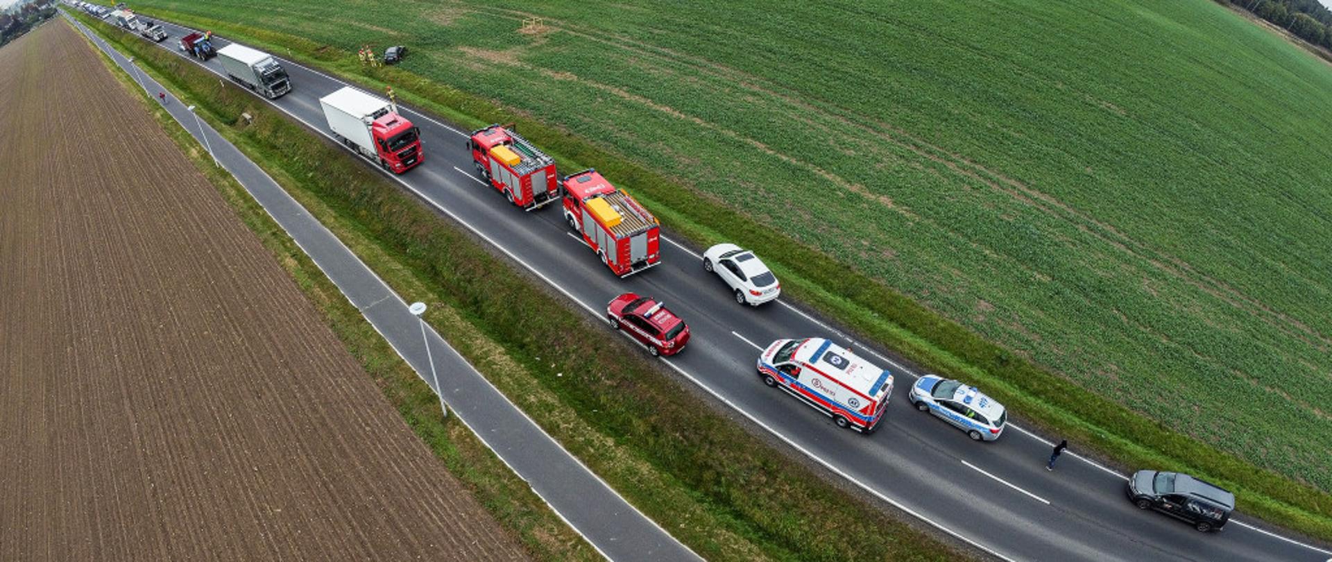 Zdjęcie z lotu ptaka. Przedstawia samochody służb ratowniczych oraz cywilne pojazdy stojące na drodze. W tle, na polu leży uszkodzony samochód osobowy.