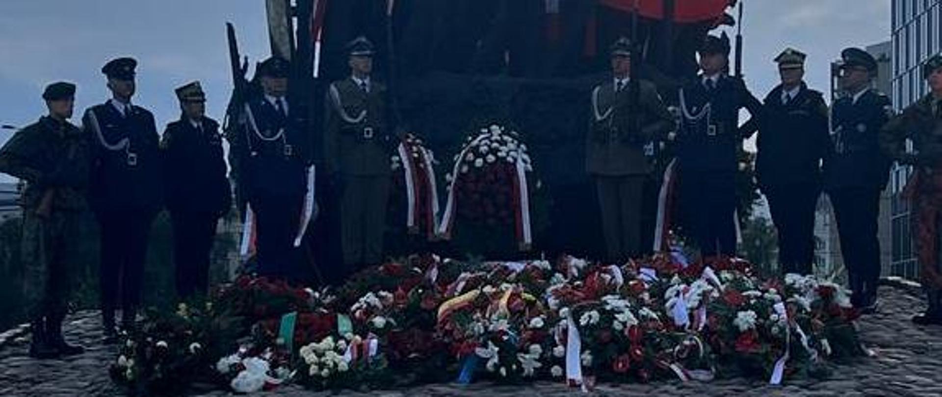 Umundurowani funkcjonariusze kilku formacji w tym wojskowych na tle pomnika i złożonych kwiatów