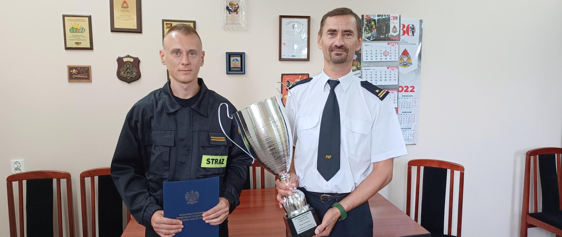 Zdjęcie przedstawia dwóch funkcjonariuszy, strażaków stojących frontem do fotografa. Jeden z nich ubrany w umundurowanie koszarowe koloru czarnego trzyma w rękach ozdobną granatową teczkę symbolizującą otrzymanie nagrody za osiągnięte sukcesy w sporcie. Drugi funkcjonariusz ubrany w umundurowanie galowe, biała koszulę na krótki rękaw oraz ciemno-granatowe spodnie trzyma w rękach duży, połyskujący srebrny (metalowy) puchar. W tle widać drewniany stół z krzesłami oraz ścianę, na której wiszą nagrody i podziękowania dla strażaków komendy powiatowej PSP w Aleksandrowie Kujawskim. Uroczystość odbywa się w gabinecie komendanta powiatowego Państwowej Straży Pożarnej.