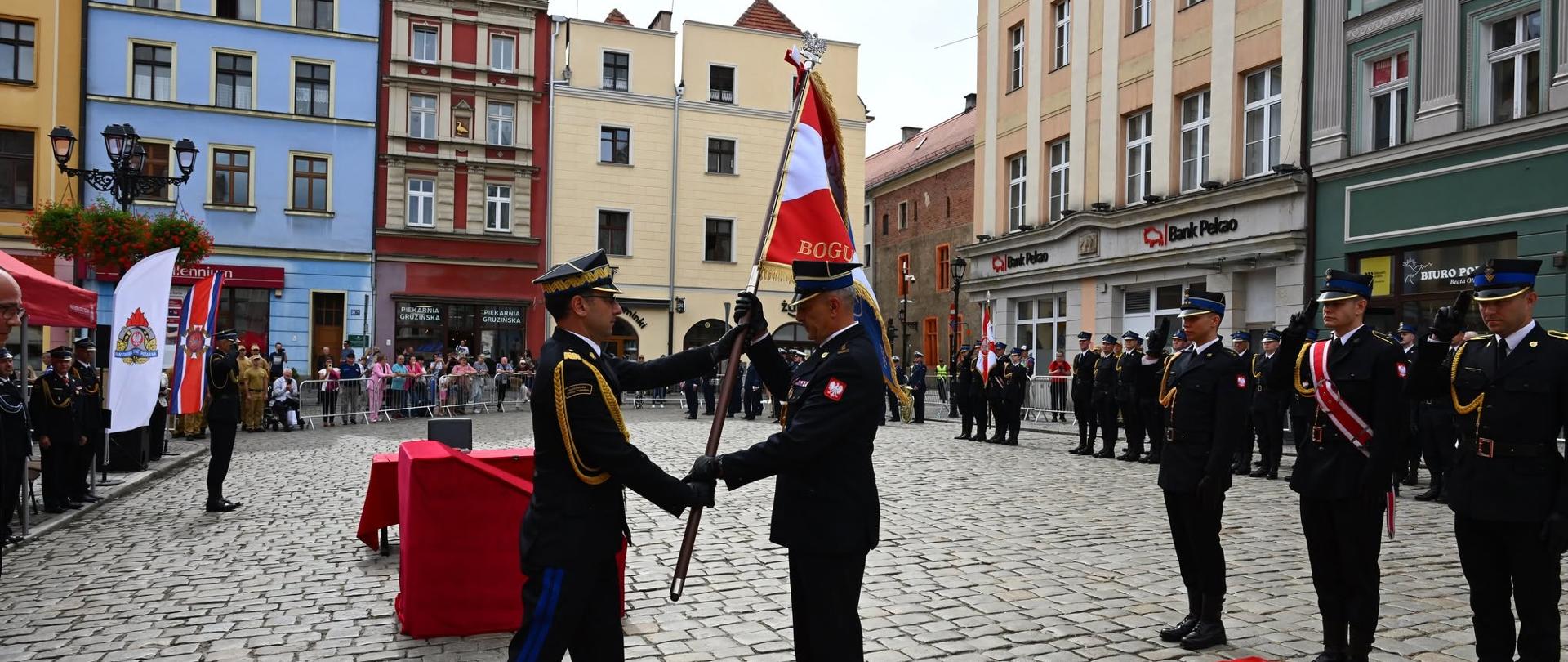 Dwóch funkcjonariuszy przekazuje sobie sztandar. W centrum miasta.