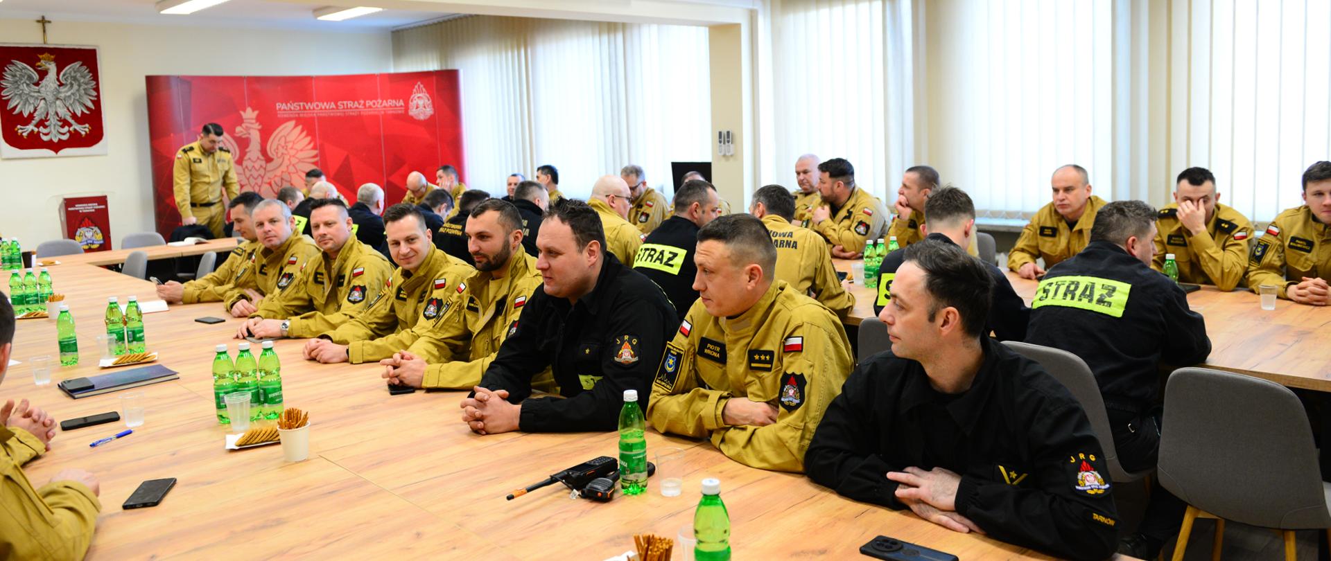 Na zdjęciu widać grupę strażaków siedzących przy długich stołach w sali konferencyjnej. Część z nich nosi beżowe mundury, a inni czarne uniformy z oznaczeniami "STRAŻ". Na stołach znajdują się napoje, przekąski oraz notatniki. W tle widać czerwoną ściankę z logo Państwowej Straży Pożarnej, a na ścianie wisi godło Polski. Strażacy wydają się uczestniczyć w szkoleniu.