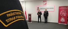 Zdjęcie przedstawia scenę z uroczystej zbiórki z okazji Narodowego Święta Niepodległości, która odbyła się w Sali Sztabowej Komendy Miejskiej Państwowej Straży Pożarnej w Mysłowicach. Ujęcie jest wykonane z perspektywy uczestnika stojącego w szeregu, patrzącego w kierunku sceny.
Pierwszy Plan: Na pierwszym planie, w lewym dolnym rogu, widoczne jest ramię i część pleców osoby w ciemnym, galowym mundurze, z naszywką na rękawie, na której złotymi nićmi wyhaftowany jest napis "PAŃSTWOWA STRAŻ POŻARNA". To element kadru wprowadza widza w uroczystą atmosferę wydarzenia służbowego.
Scena i Kierownictwo: W centrum zdjęcia, na podwyższeniu lub przed ścianą, stoją dwaj mężczyźni w ciemnych, galowych mundurach PSP, którzy prawdopodobnie pełnią funkcje dowódcze i przewodniczą uroczystości. Jeden z nich stoi przy mównicy.
Tło i Symbole: Tło jest jasne, w kolorach bieli i szarości. Na ścianie wiszą dwa główne elementy:
Centralny Panel: Czerwono-biały panel z napisem "PAŃSTWOWA STRAŻ POŻARNA KOMENDA MIEJSKA PSP W MYSŁOWICACH".
Banner Okolicznościowy: Po prawej stronie widać duży, czerwony banner z logo KSRG i napisem "30 LAT".
Symbole Narodowe: Nad centralnym panelem znajduje się godło Polski, a po lewej stronie sceny widoczna jest flaga Polski na stojaku.
Pozostali Uczestnicy: Po prawej stronie widać część kolejnych uczestników zbiórki (w tym kobietę i mężczyznę w galowych mundurach), którzy oczekują na wręczenie awansów na wyższe stopnie służbowe, powierzenia na nowe stanowiska lub medali okolicznościowych z okazji 30-lecia KSRG.
Zdjęcie doskonale oddaje podniosły i uroczysty charakter zbiórki związanej ze świętem państwowym i podsumowującej osiągnięcia służbowe.
