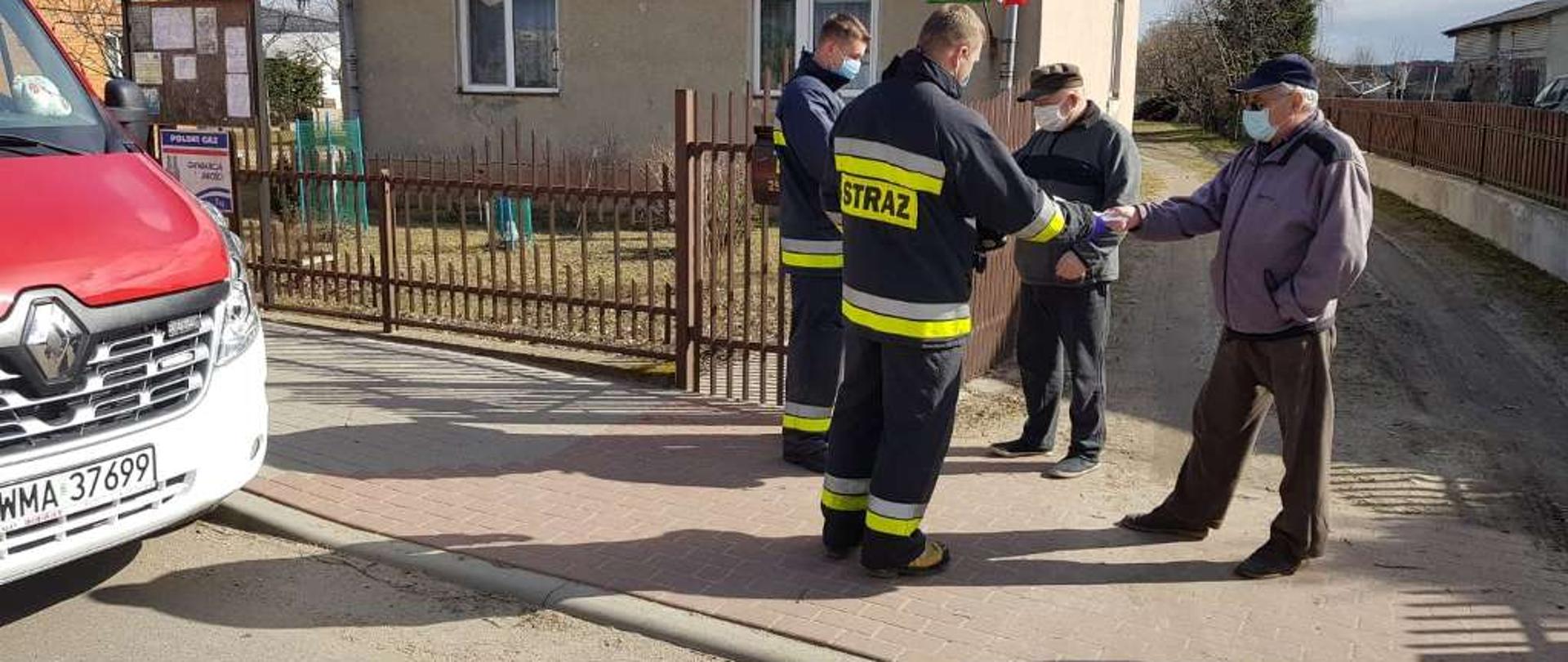 Dwóch strażaków wręcza ulotki dwóm starszym mężczyznom. Po lewej stronie widoczna przednia część samochodu strażackiego w kolorze czerwonym
